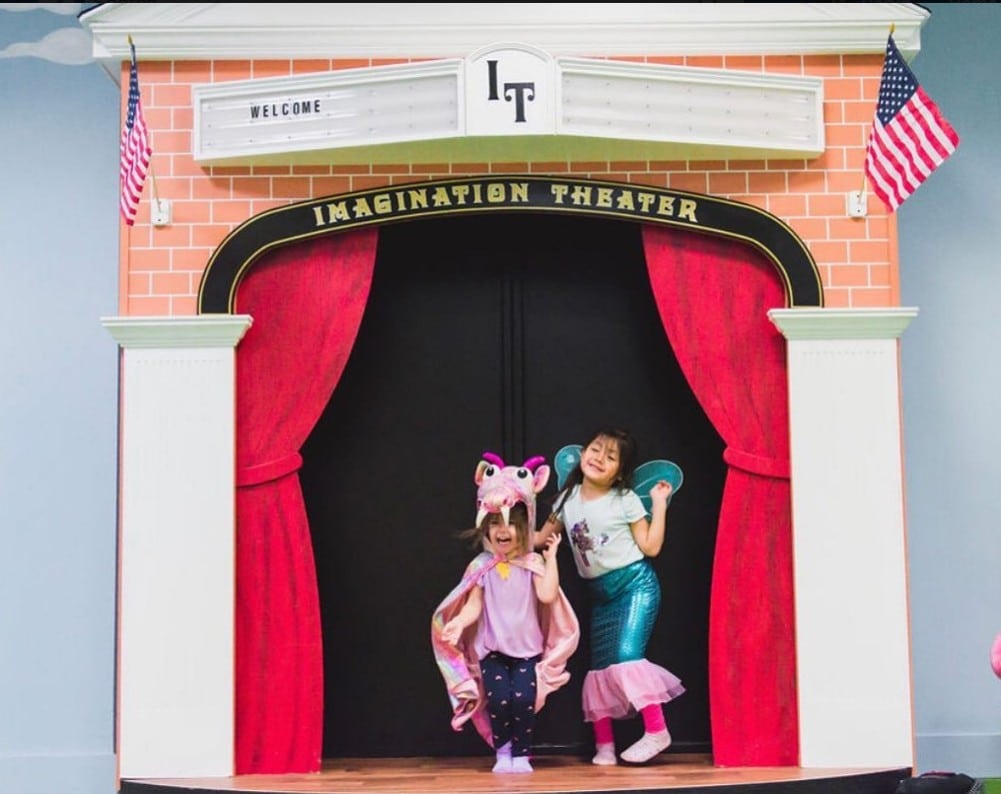 Two children in costumes pose on the Main Street Stage playhouse, featuring red curtains, a brick facade, and two American flags.