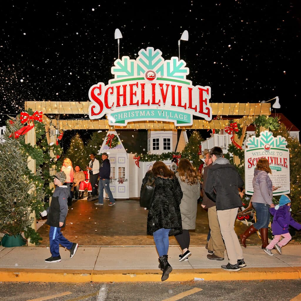 People walk into Schellville Christmas Village at night, passing festive decorations and a welcome sign, with artificial snow falling.
